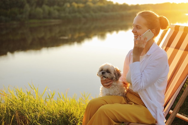 Ritratto di vista laterale di felice donna positiva che indossa una camicia bianca seduta con il suo cane pechinese vicino al fiume sulla sedia a sdraio e parlando al telefono cellulare con una piacevole conversazione