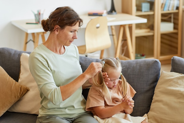 Side view portrait of loving mother combing hair of cute girl with down syndrome and tying it in pigtails, copy space