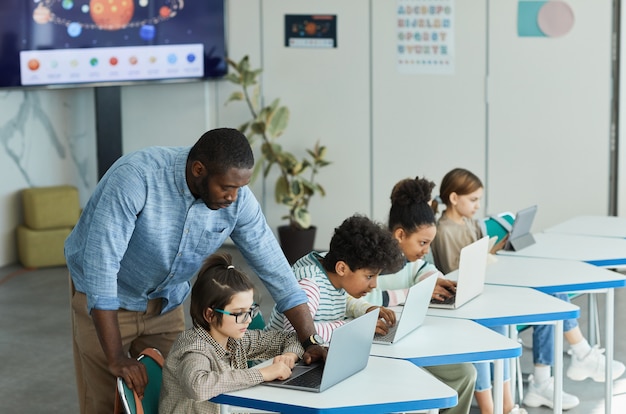 Side view portrait of male teacher helping children using computers in IT class, copy space