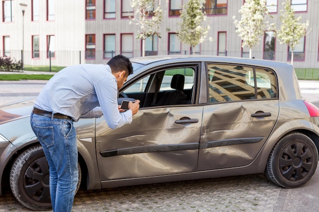 Foto ritratto di vista laterale di un uomo che indossa jeans e maglietta che fa foto di un'auto danneggiata dopo un incidente automobilistico fotografando ammaccature e graffi sulle portiere del veicolo e sul parafango scatto all'aperto