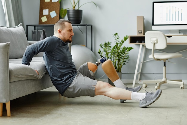 Side view portrait of man with prosthetic leg doing exercises\
at home in minimal interior
