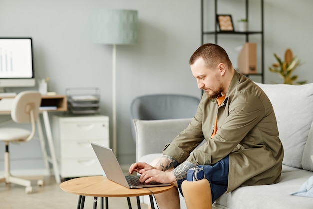 Side view portrait of man with prosthetic leg using laptop while working in minimal office interior