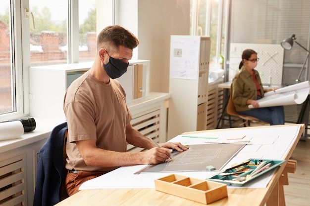 Photo side view portrait of mature bearded architect wearing mask while sitting at drawing desk in sunlight,