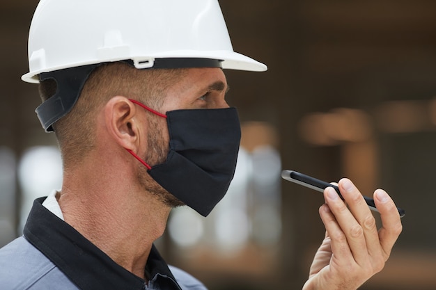 Side view portrait of mature worker wearing mask and recording voice message while working on industrial site, 