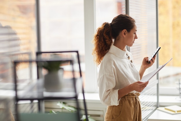 Ritratto di vista laterale della moderna imprenditrice di successo tenendo lo smartphone mentre in piedi dalla finestra in ufficio, copia dello spazio