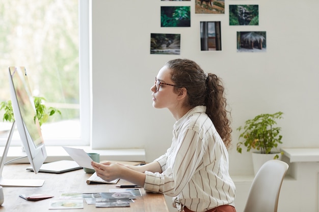 白いオフィス、コピースペースでPCで作業しながら写真を保持している現代の若い女性の側面図の肖像画