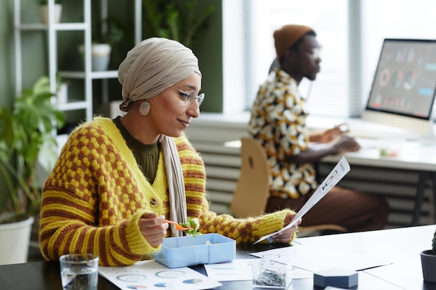 Side view portrait of muslim young woman wearing headscarf and eating healthy lunch in office