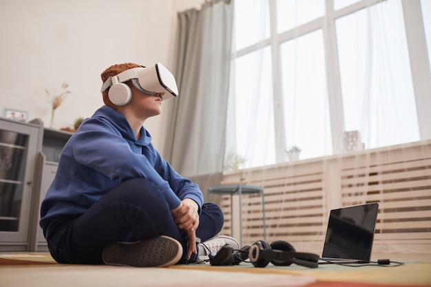 측면 보기 초상화 또는 VR 헤드셋을 착용한 빨간 머리 10대 소년이 바닥에 앉아 비디오 게임을 하는 동안 공간을 복사합니다.