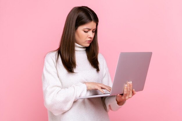 Side view portrait of pensive girl works online looks at laptop display with thoughtful expression wearing white casual style sweater Indoor studio shot isolated on pink background
