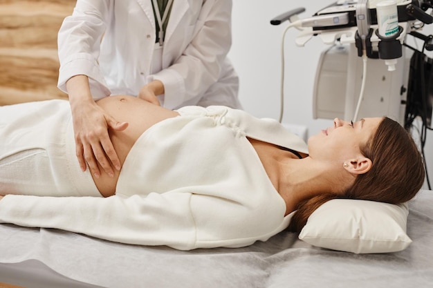 Side view portrait of pregnant young woman preparing for ultrasound in medical clinic with doctor ex...