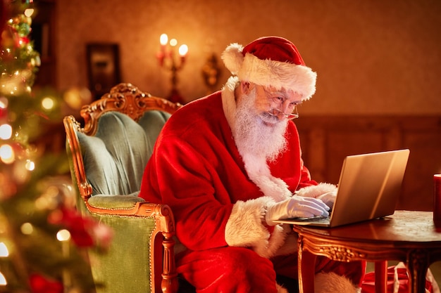 Side view portrait of santa claus using laptop by christmas tree and smiling copy space