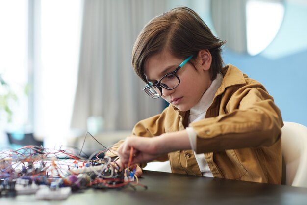 Side view portrait of schoolboy working with electric wires while building robot in engineering class