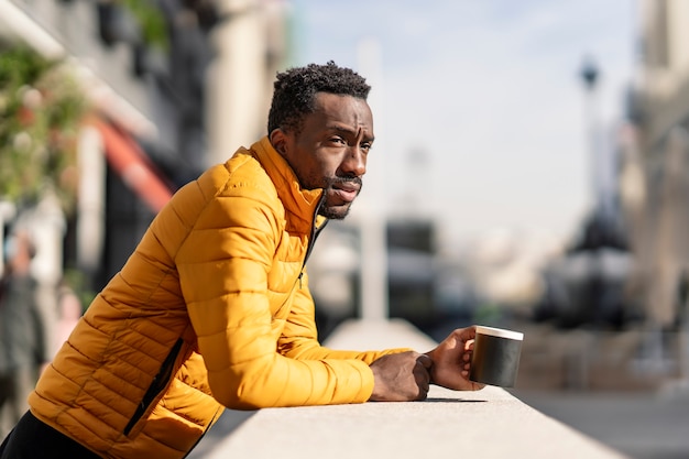 Ritratto di vista laterale di un uomo africano serio che si appoggia su un balcone che tiene una tazza di caffè contemplando le viste in una città