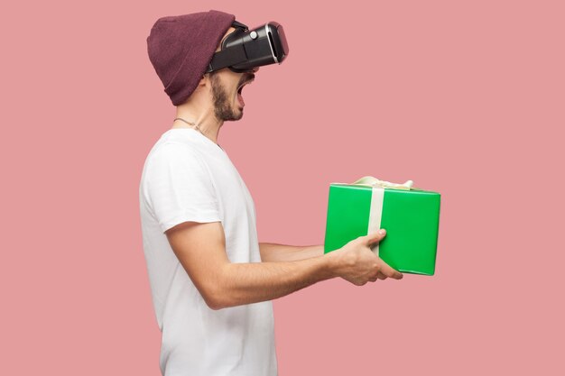 白いシャツ、カジュアルな帽子の立っている、vrを身に着けている、口を開けて緑のプレゼントボックスを保持しているショックを受けたひげを生やした若いヒップスターの側面図の肖像画。屋内、孤立した、スタジオショット、ピンクの背景