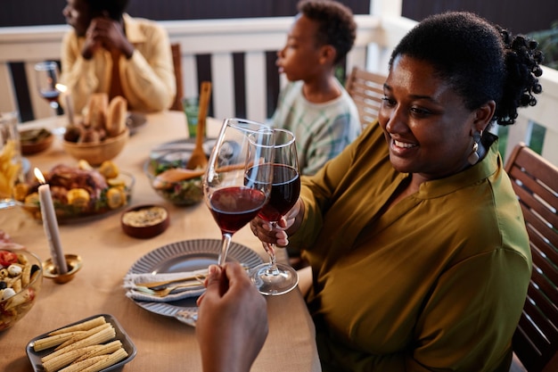 Side view portrait of smiling black woman clinking wine glasses at dinner table outdoors in cozy set