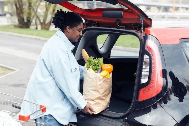 Side view portrait of smiling black woman putting groceries in car trunk in parking lot copy space