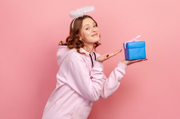 Side view portrait of smiling curly haired teenage girl in hoodie and halo on head holding blue wrapped gift box, holiday greeting. Indoor studio shot isolated on pink background