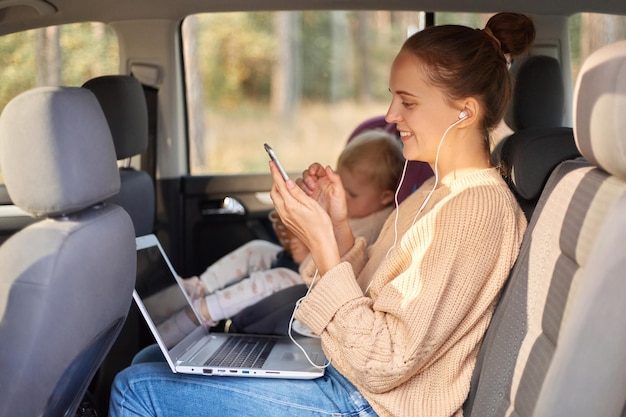 Side view portrait of smiling extremely woman with earphones listening musing from smartphone working on laptop while sitting with her baby daughter in safety chair on backseat of the auto