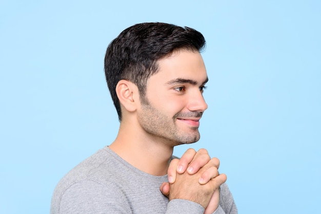 Side view portrait of a smiling handsome Caucasian man holding his hands together wishing with eyes open in isolated studio blue background
