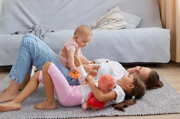 Side view portrait of smiling optimistic woman lying on floor with her daughters baby kid sitting on moms belly elder child lying near mommy family spending happy time together