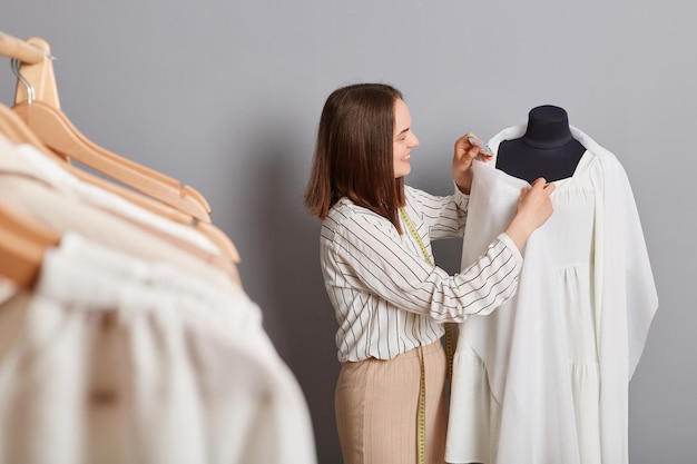 side view portrait of smiling positive female sewer standing near mannequin with new dress isolated over gray background using pins working on new collection with pleasure