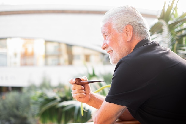 Side View Portrait of Smiling Senior Bearded Man Smoking Pipe Outdoor On the Home Balcony