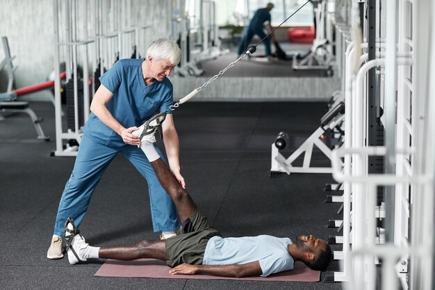 Side view portrait of smiling senior therapist helping young man with rehabilitation exercises in gy