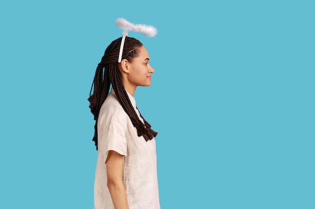 Side view portrait of smiling woman with black dreadlocks with nimbus over her head, looking ahead with toothy smile, expressing positive emotions. Indoor studio shot isolated on blue background.