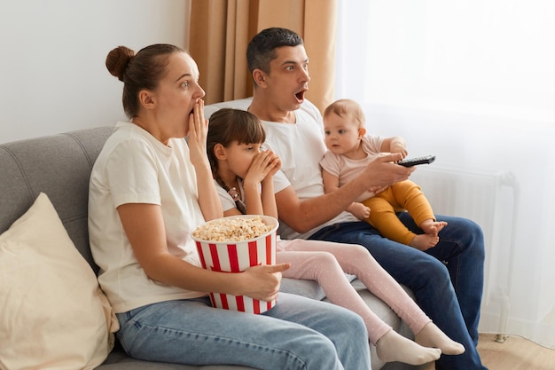 Side view portrait of surprised family sitting on sofa and\
watching horror movie with popcorn expressing shocked emotions\
while watching tv together spending leisure time together