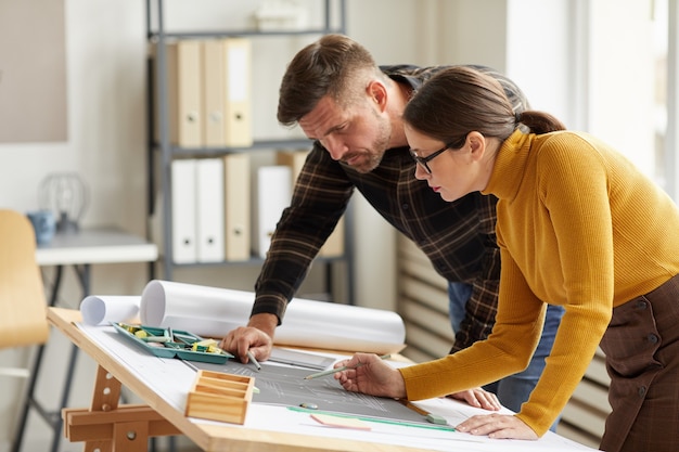 Side view portrait of two architects pointing at floor plan while working on blueprints at workplace, 