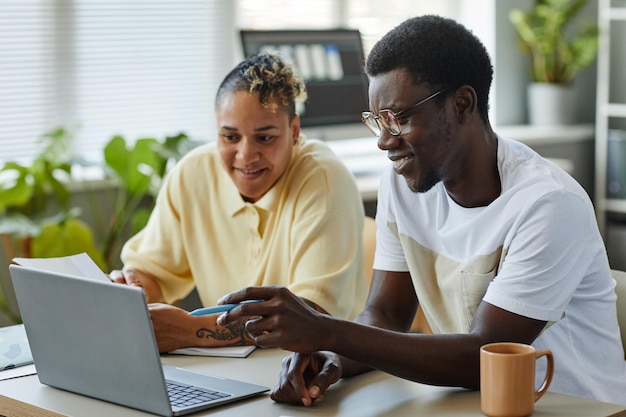 Ritratto di vista laterale di due persone di colore sorridenti che usano il laptop insieme in ufficio e indossano abiti casual