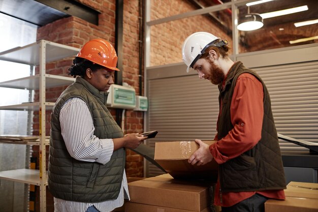 Side view portrait of two workers wearing hardhats counting boxes in factory warehouse and using tab