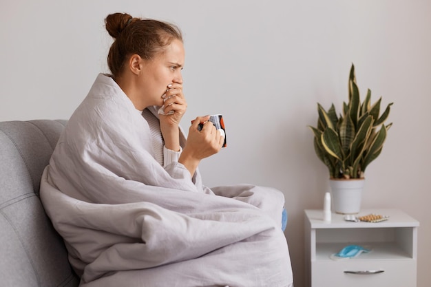 Ritratto di vista laterale di donna malsana seduta su tosse e tosse, bere tè, soffrire di influenza, avere febbre alta e mal di testa, assistenza sanitaria.