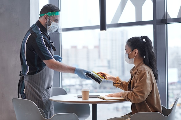 Вид сбоку портрет официанта в маске и перчатках, обслуживающего клиентку в кафе с мерами безопасности covid