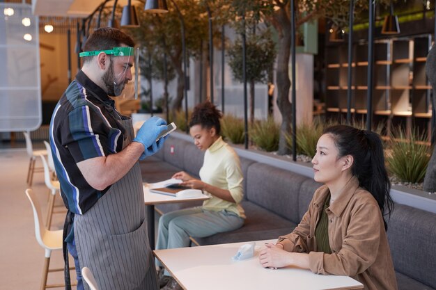 Вид сбоку портрет официанта в маске и перчатках во время принятия заказа в кафе с мерами безопасности covid, копией пространства