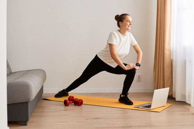 Side view portrait of woman doing fitness at home watching fitness videos on Internet and doing stretching workout looking away with positive expression using laptop