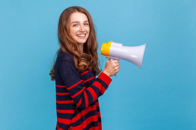 Side view portrait of woman wearing striped casual style sweater holding megaphone in hands looking at camera with happy expression and toothy smile Indoor studio shot isolated on blue background