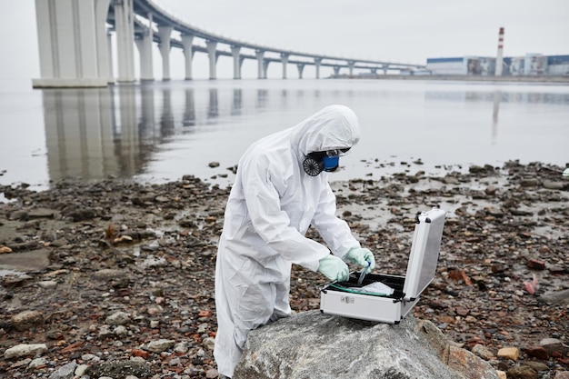 Side view portrait of worker wearing hazmat suit collecting probes by water toxic waste and pollutio...