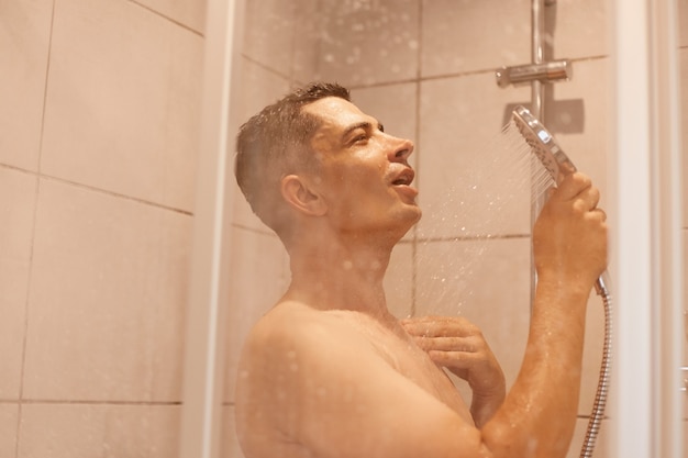 Photo side view portrait of young adult brunette man taking hot and cold water, refreshing himself after early waking up, having satisfied look, posing naked in bathroom.