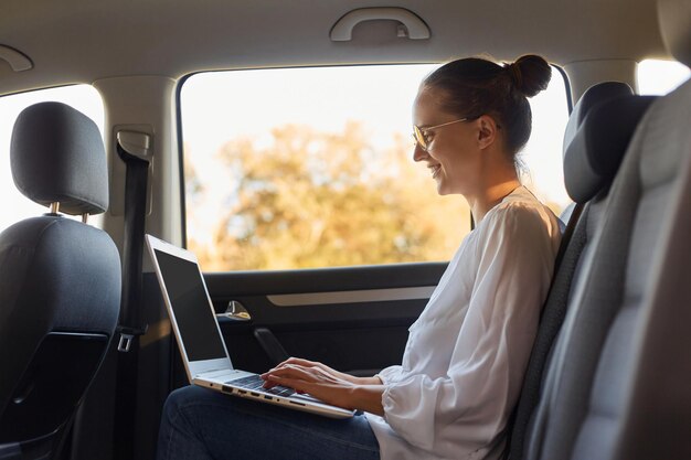 Side view portrait of young adult Caucasian beautiful businesswoman using laptop while sitting on a backseat of a car looking on display of notebook with toothy smile