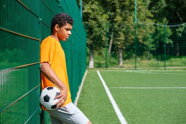 Vista laterale ritratto di giovane uomo afro-americano che tiene in mano un pallone da calcio mentre in piedi con la schiena contro la recinzione in un campo sportivo all'aperto, copia spazio