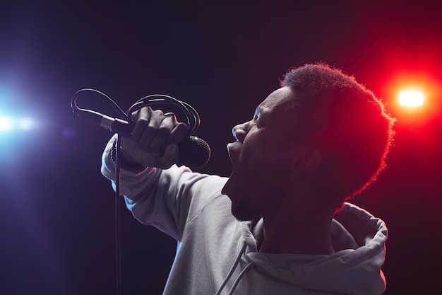 Photo side view portrait of young african-american man singing to microphone emotionally while standing on stage in lights