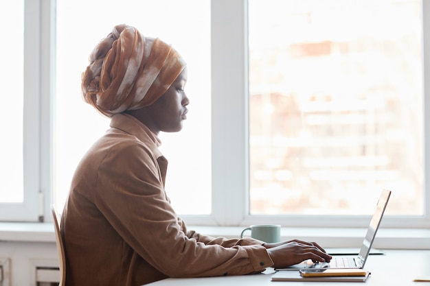 사무실 창가에 앉아 컴퓨터를 사용하는 동안 머리 스카프를 두른 젊은 아프리카계 미국인 여성의 측면 초상화, 복사 공간