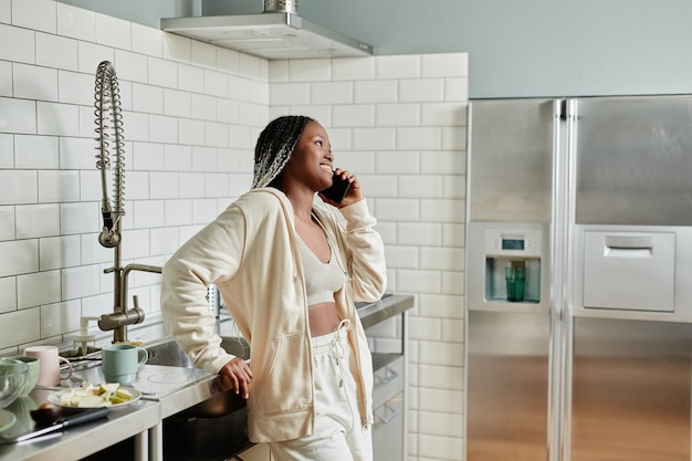 Портрет молодой афроамериканской женщины, разговаривающей по смартфону, стоя на кухне, вид сбоку ...