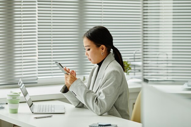 Side view portrait of young Asian businesswoman using smartphone at desk in office interior, copy space