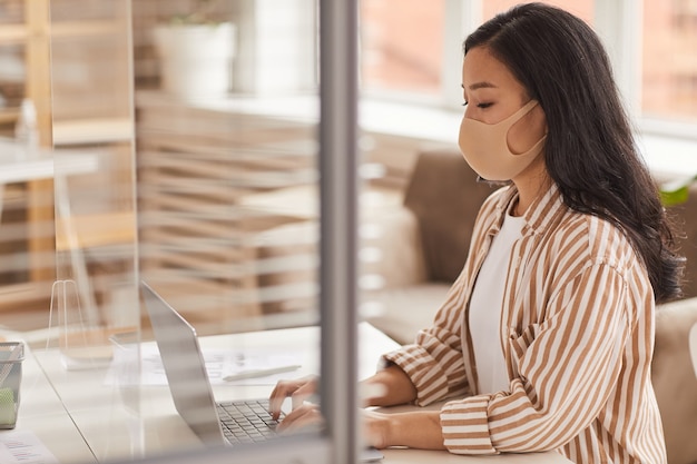 Side view portrait of young Asian woman wearing mask and using laptop while working at desk in office cubicle, copy space