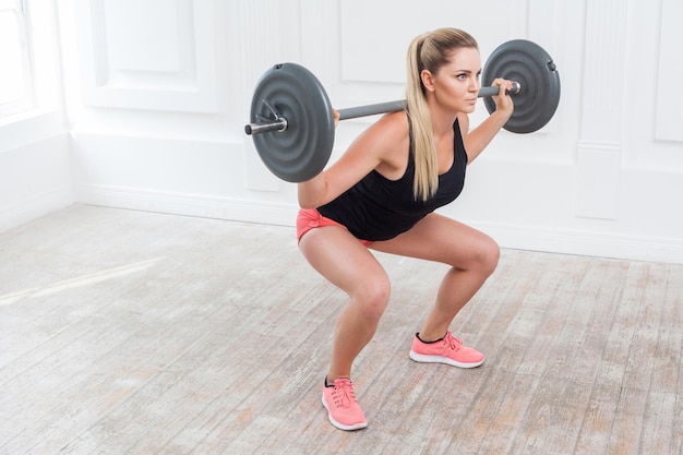 Side view portrait of young athletic beautiful bodybuilder woman in pink shorts and black top doing squats and exercising at the gym with the barbell on white wall. indoor studio shot.