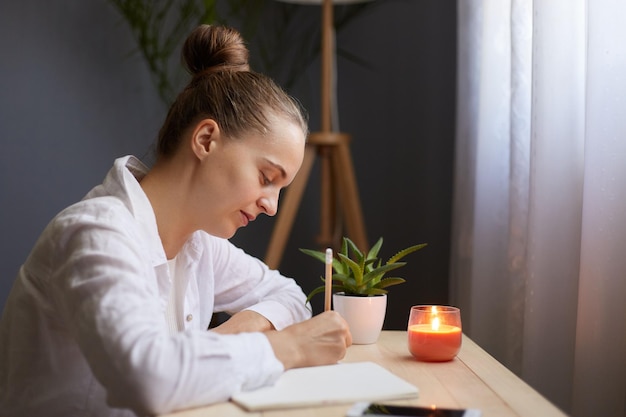 Side view portrait of young beautiful focused woman writing down notes while sitting at table in office in front of the window working or studying female with bun hairstyle wearing whie shirt