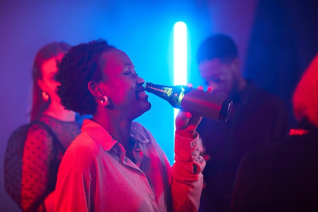 Side view portrait of young black woman enjoying drinks at party in neon light copy space