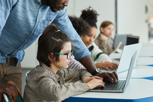 Photo side view portrait of young boy using computer in it class with teacher helping him